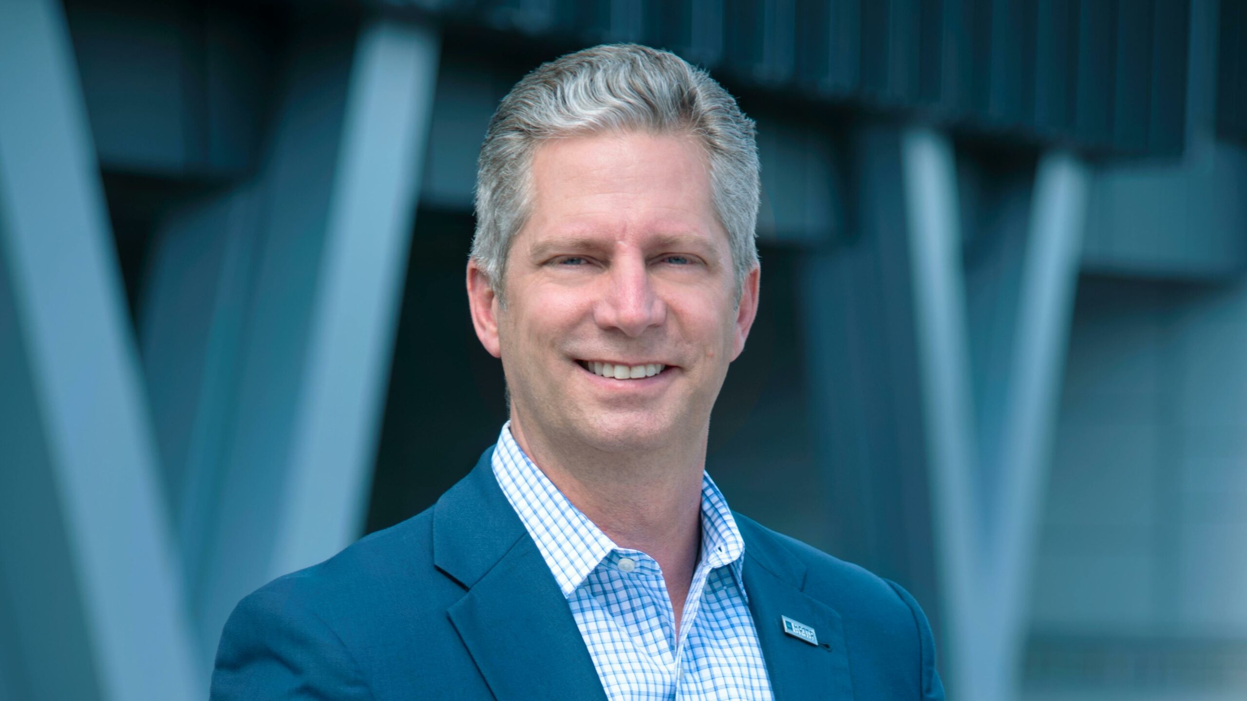 A smiling man with short gray hair wearing a blue blazer and a checkered shirt stands outdoors in front of a modern building with large vertical beams.