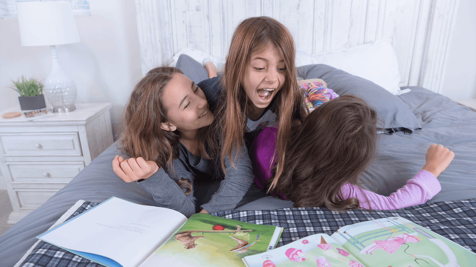 Three young girls are laughing and playing on a bed with several open picture books in front of them. The room is bright and cozy, with a nightstand, lamp, and potted plant visible in the background.