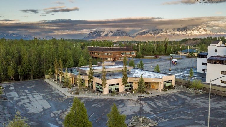 Aerial view of a mostly empty parking lot surrounding a modern building with large windows, set against a backdrop of dense green trees and snow-capped mountains under a cloudy sky.