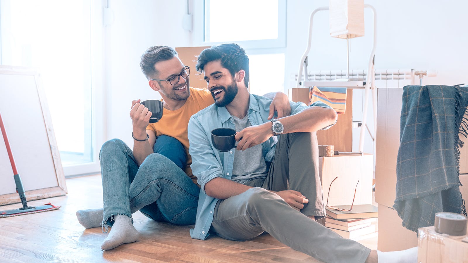 Two men sit on the floor among moving boxes, smiling and holding coffee mugs. One wears glasses and a yellow shirt, the other a blue shirt. The bright room has sunlight streaming in and various unpacked items around them.