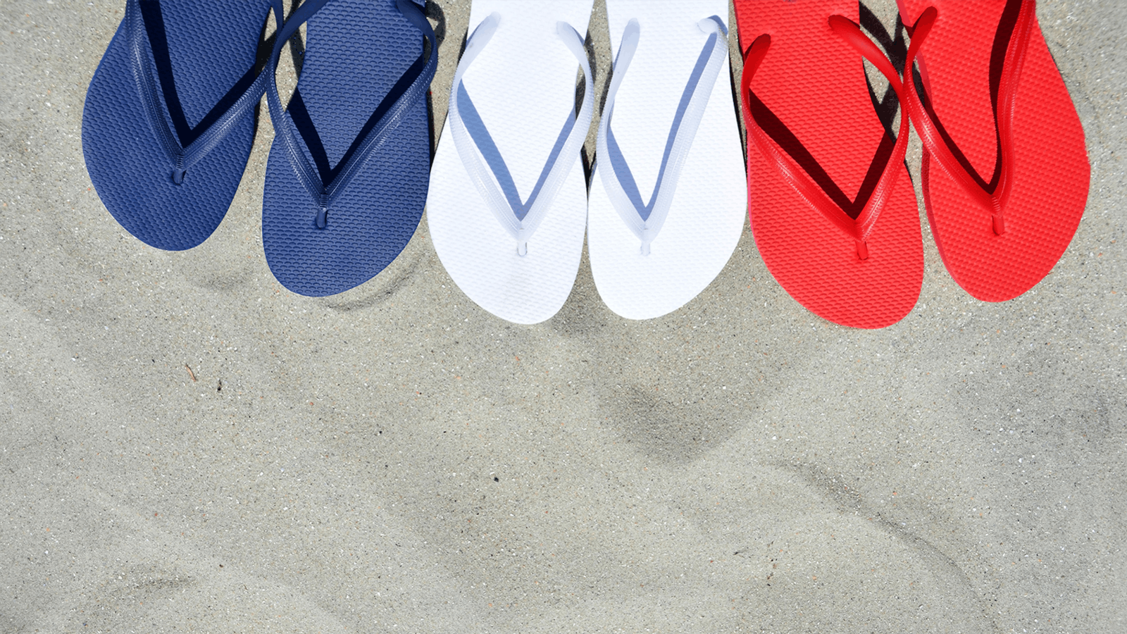Three pairs of flip-flops—one navy blue, one white, and one red—are lined up on light sandy beach, viewed from above.