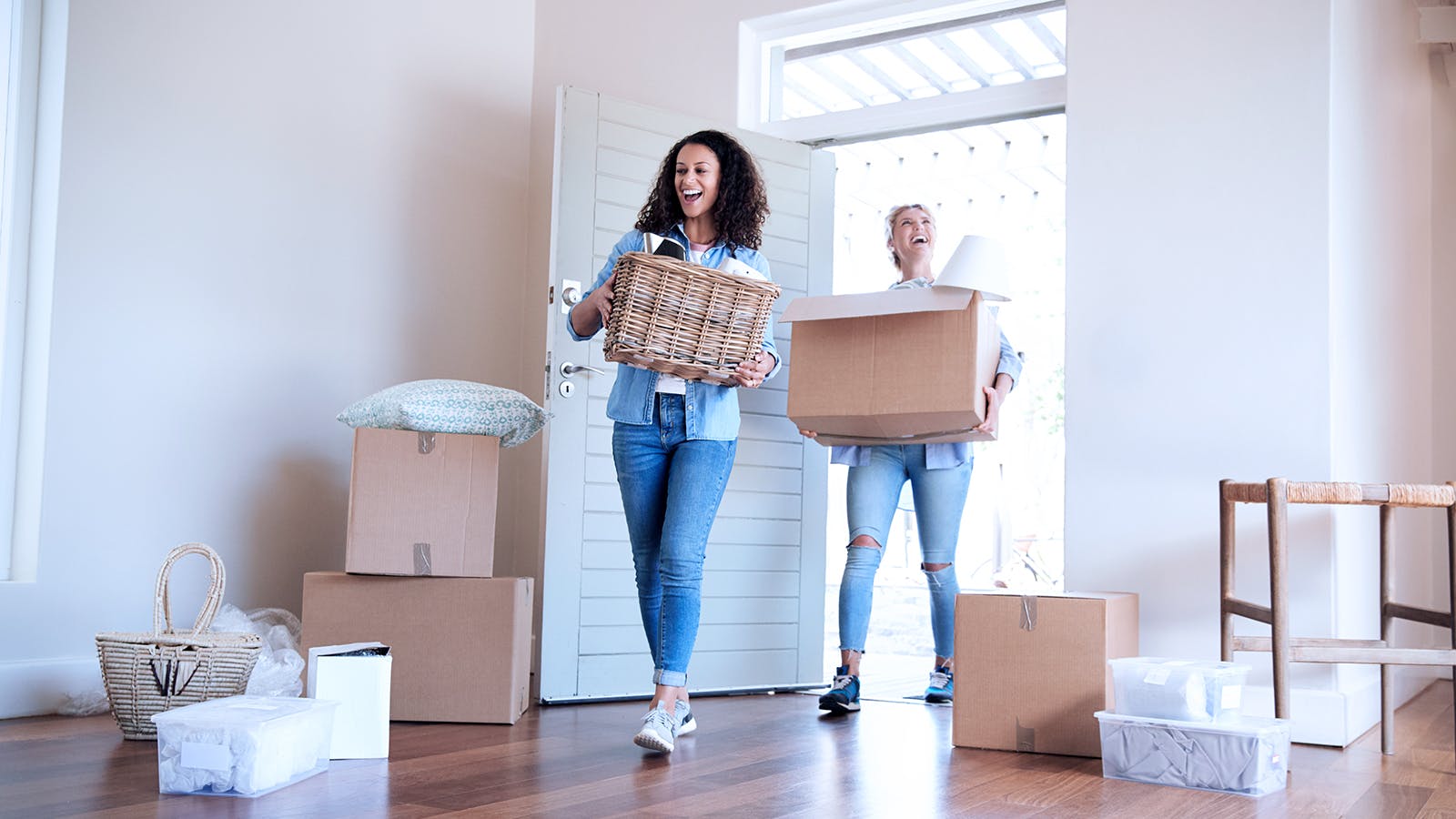 Two women smiling and carrying boxes into a bright, unfurnished room with an open door. Several cardboard boxes and household items are scattered on the wooden floor.