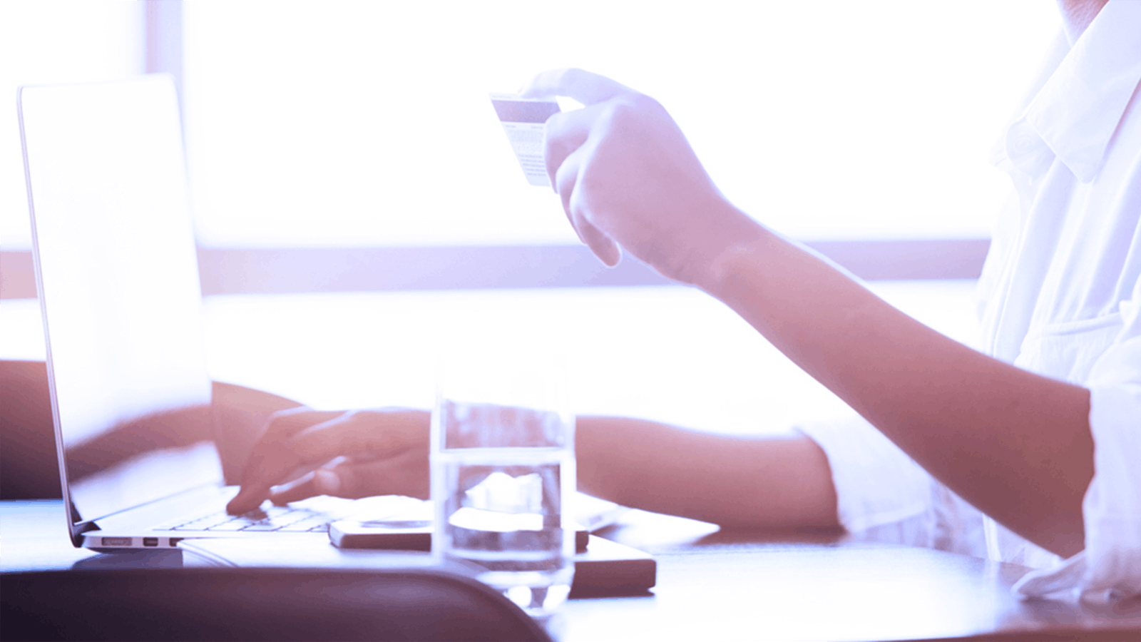 A person holds a credit card in one hand while using the other hand to type on a laptop keyboard; a glass of water and a smartphone are on the desk nearby.
