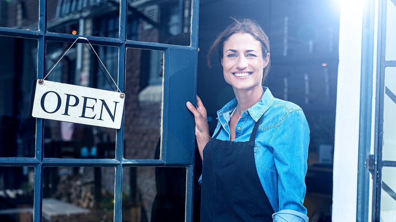 A woman wearing a denim shirt and black apron smiles while standing in the doorway of a shop with an OPEN sign hanging on the glass door. Sunlight shines in from the side.
