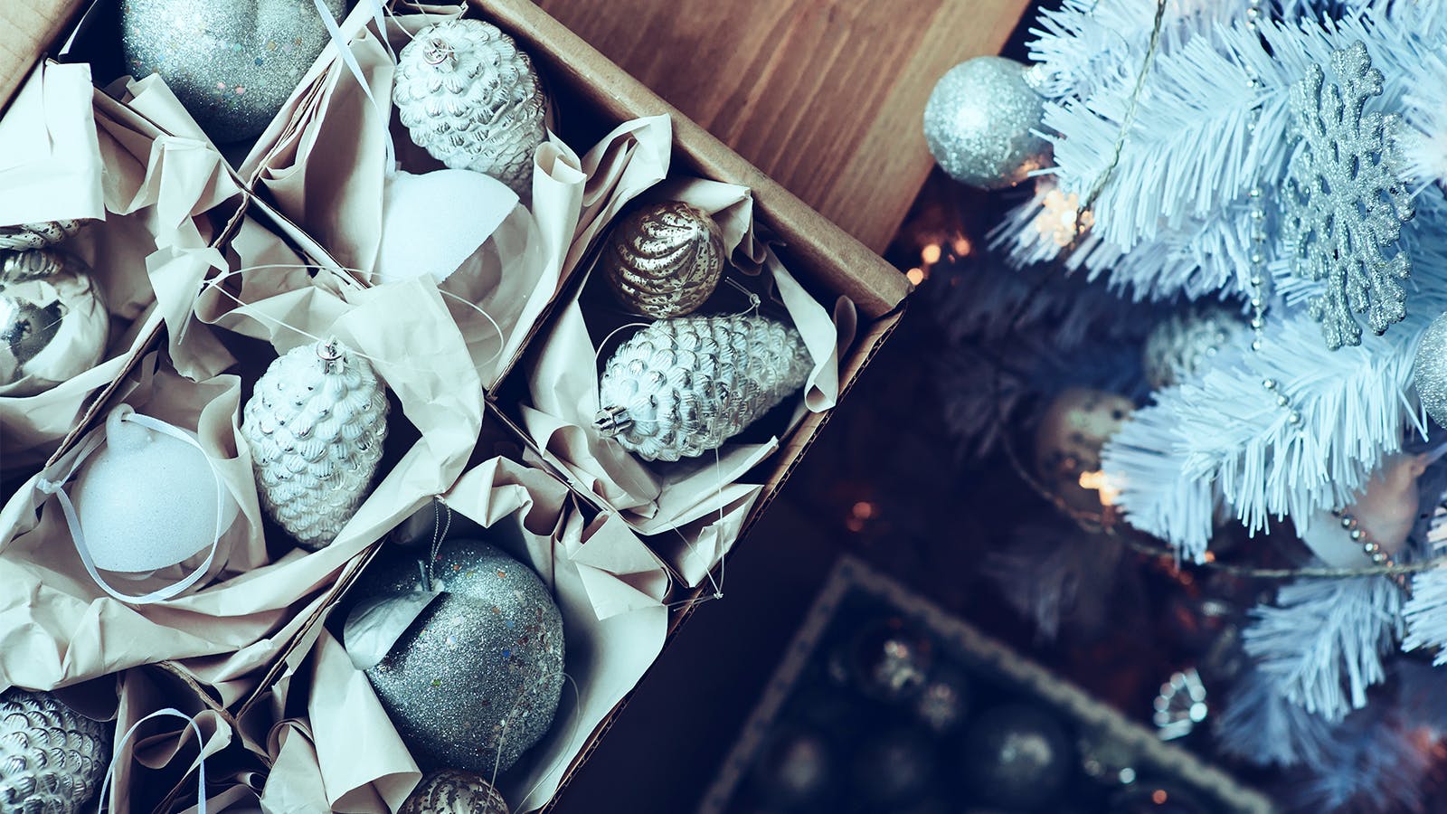 A box of silver and white Christmas ornaments, including baubles and pinecones, sits on a wooden surface next to a white Christmas tree decorated with similar ornaments.