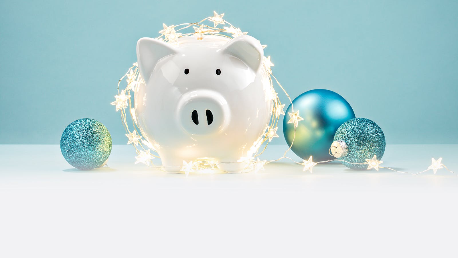 A white piggy bank is wrapped in star-shaped string lights, surrounded by several blue Christmas ornaments on a light blue background.