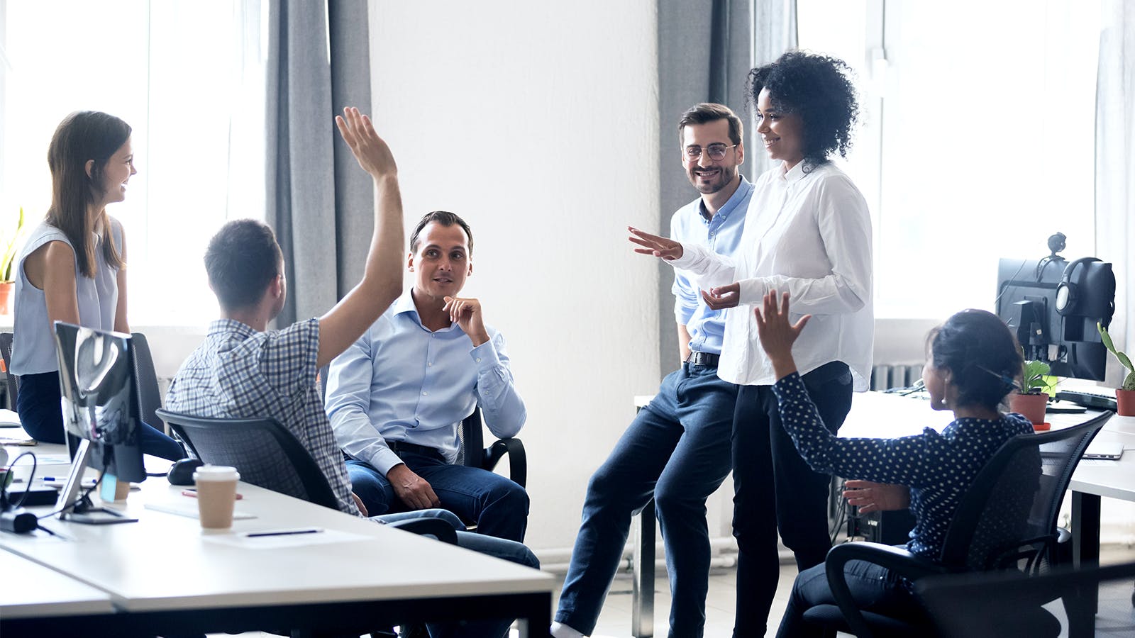 A diverse group of six colleagues in business casual attire have a lively discussion in a bright office. Two people raise their hands, and others smile or listen while sitting and standing around desks.