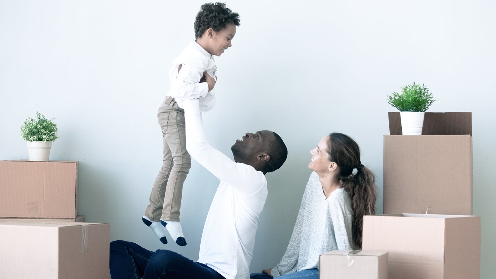 A man lifts a smiling young boy while a woman watches and smiles. They are surrounded by moving boxes and potted plants, suggesting they are moving into a new home. The background is a plain, light-colored wall.