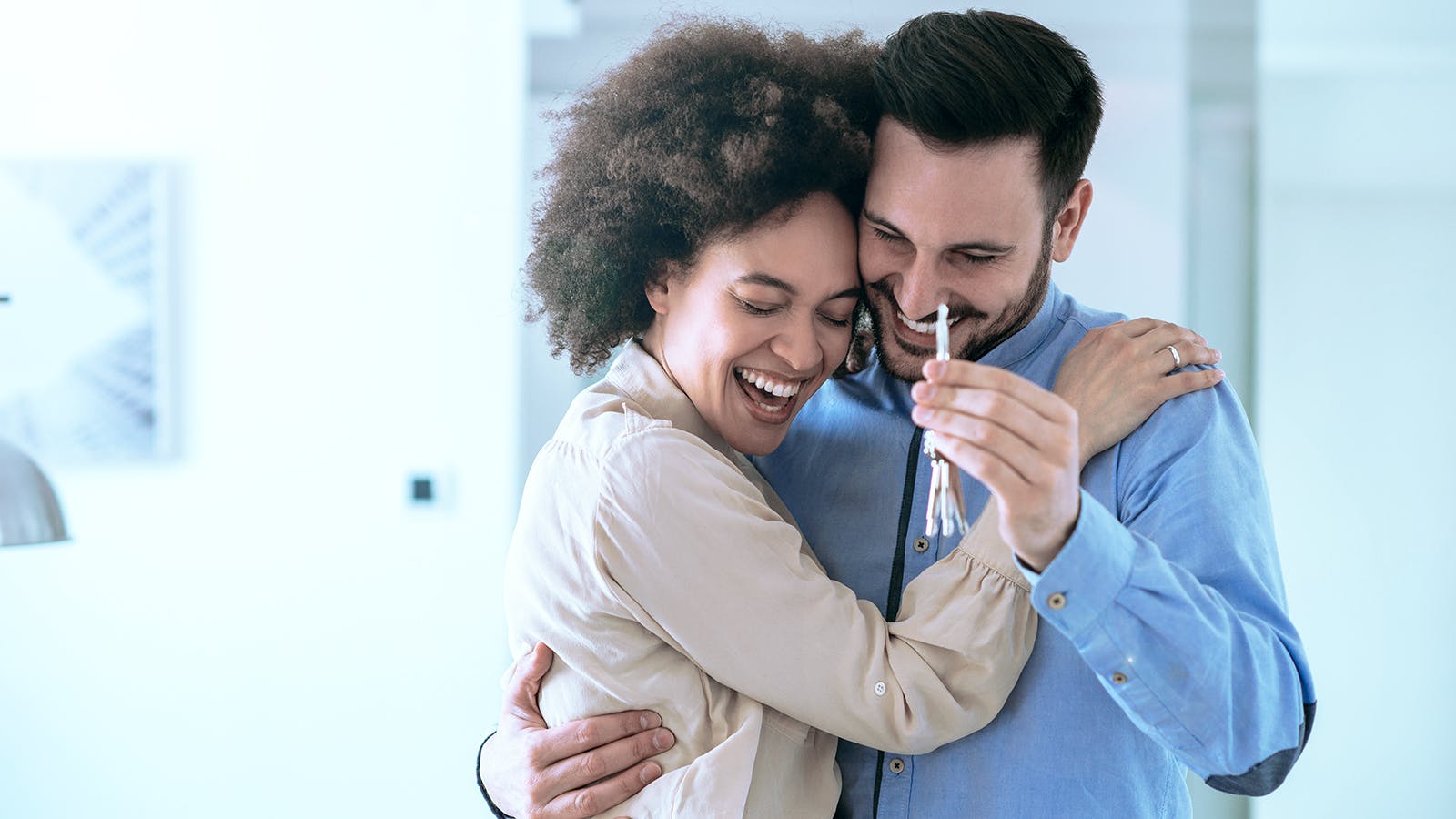 A smiling couple embraces joyfully indoors while holding up a set of house keys, suggesting they have just purchased a new home.