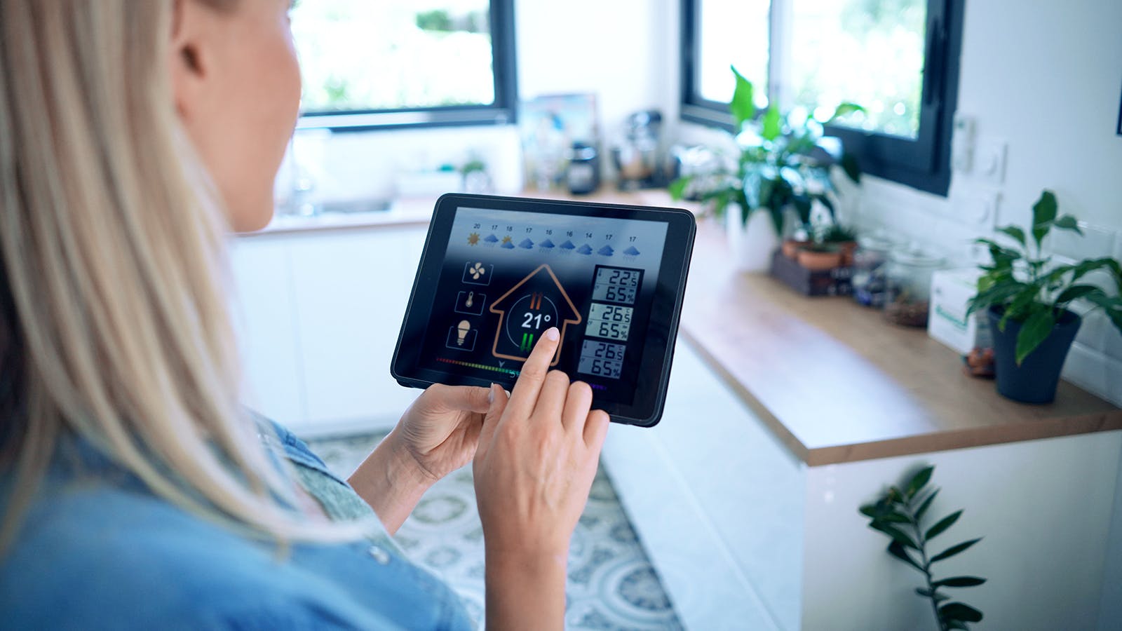 A woman stands in a modern kitchen, using a tablet to control home temperature and settings. The digital display shows various smart home options and a temperature of 21°C. Plants and bright windows are in the background.
