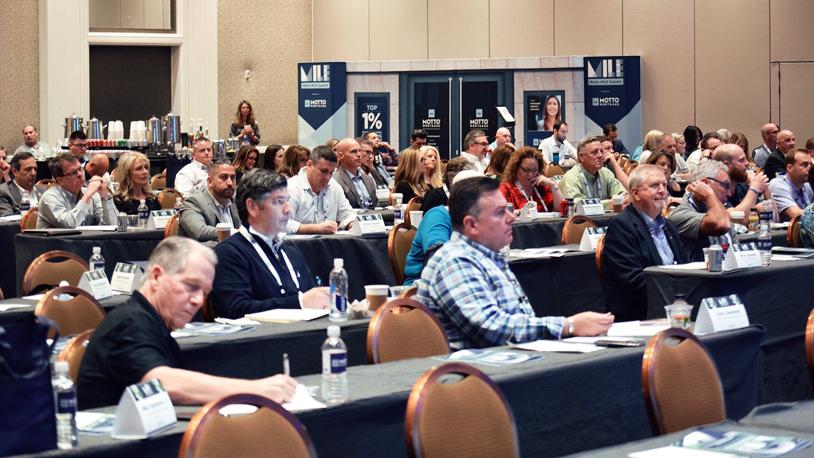 A large group of people sits at tables in a conference room, attentively listening to a presentation. Notebooks, brochures, and water bottles are on the tables. Event banners and coffee stations are visible in the background.