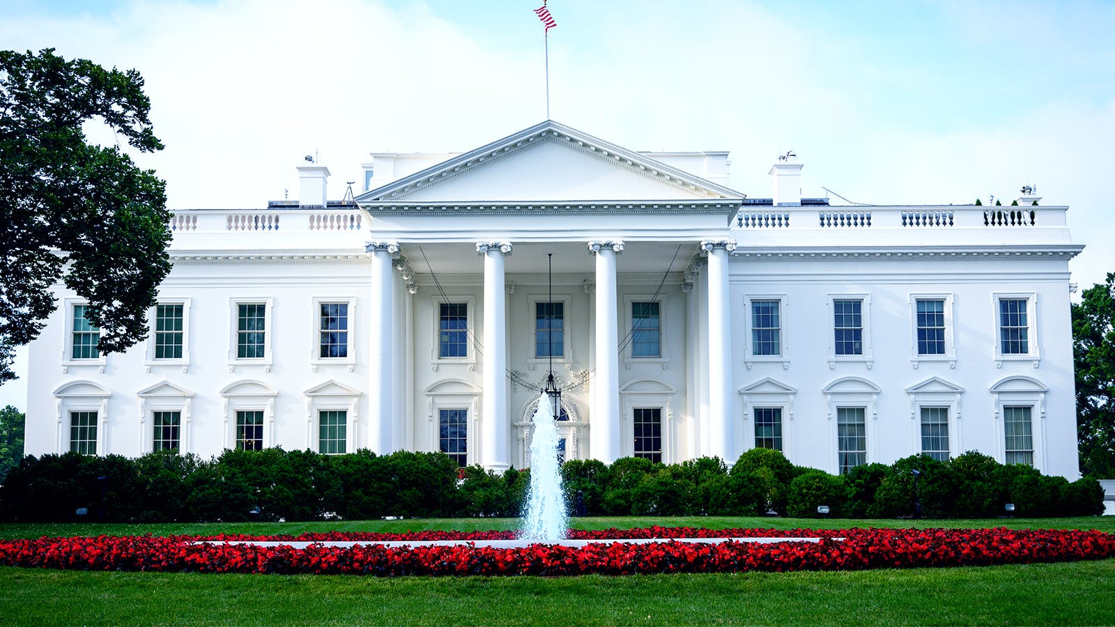 The White House, a large white neoclassical building with columns, stands behind a fountain and a garden with red flowers and green bushes under a partly cloudy sky.