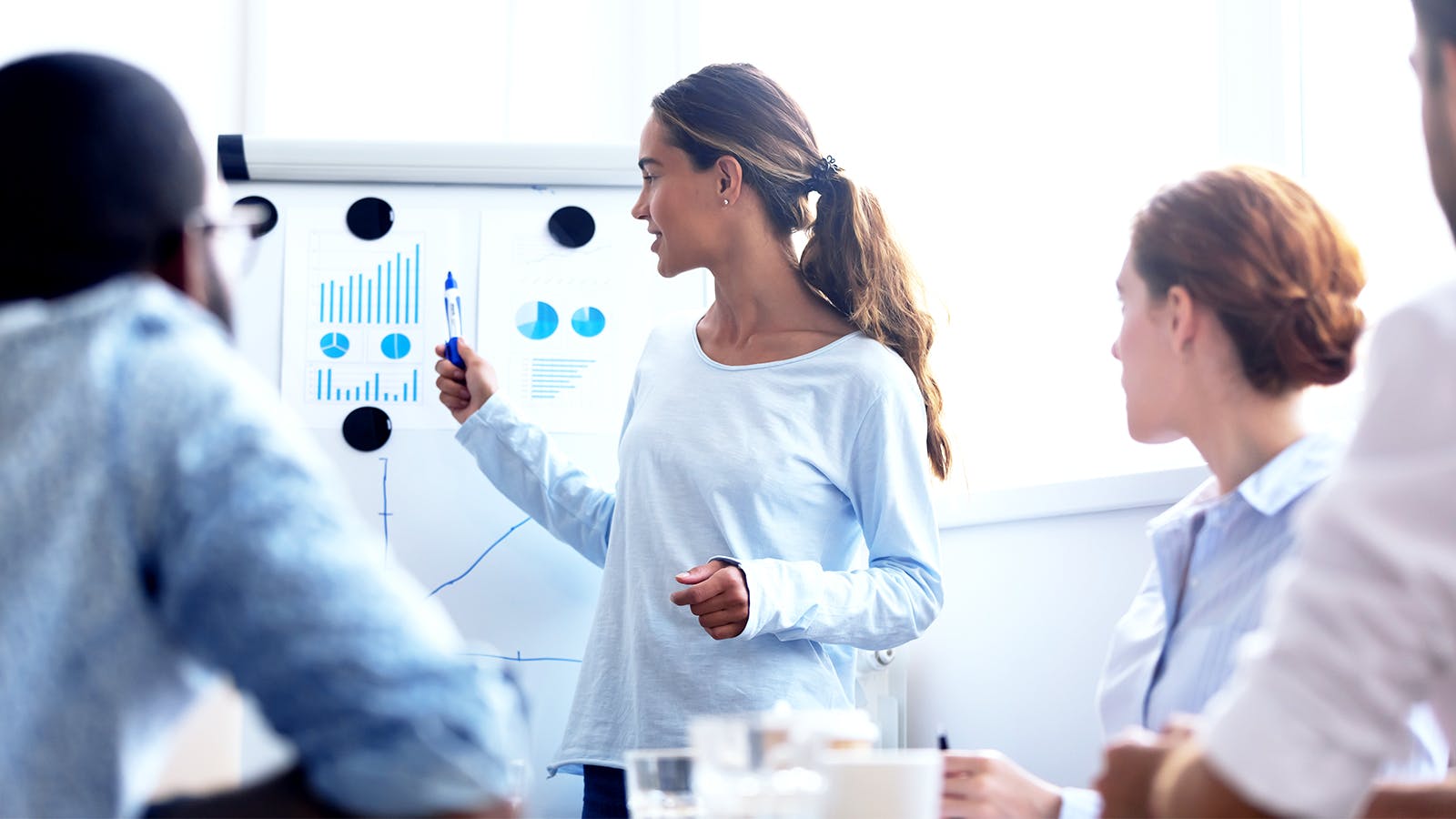 A woman stands by a flip chart presenting graphs and charts to three seated colleagues during a meeting in a bright office setting.