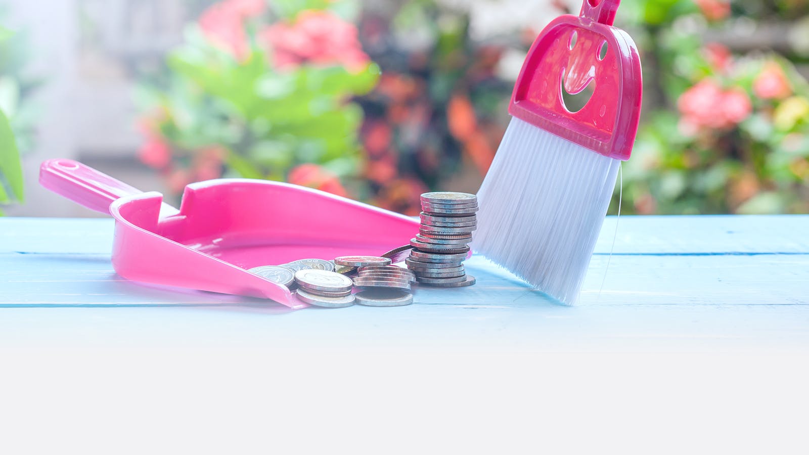 A pink dustpan and brush set on a blue surface, sweeping up stacks of coins, with blurred flowers and greenery in the background.