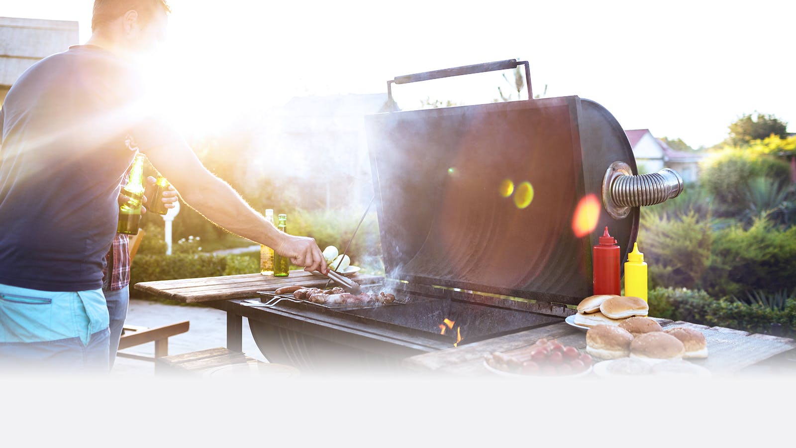A person grills food on a large outdoor barbecue while holding a drink, with buns, condiments, and vegetables on a nearby table. The scene is sunny, with bright light and green plants in the background.