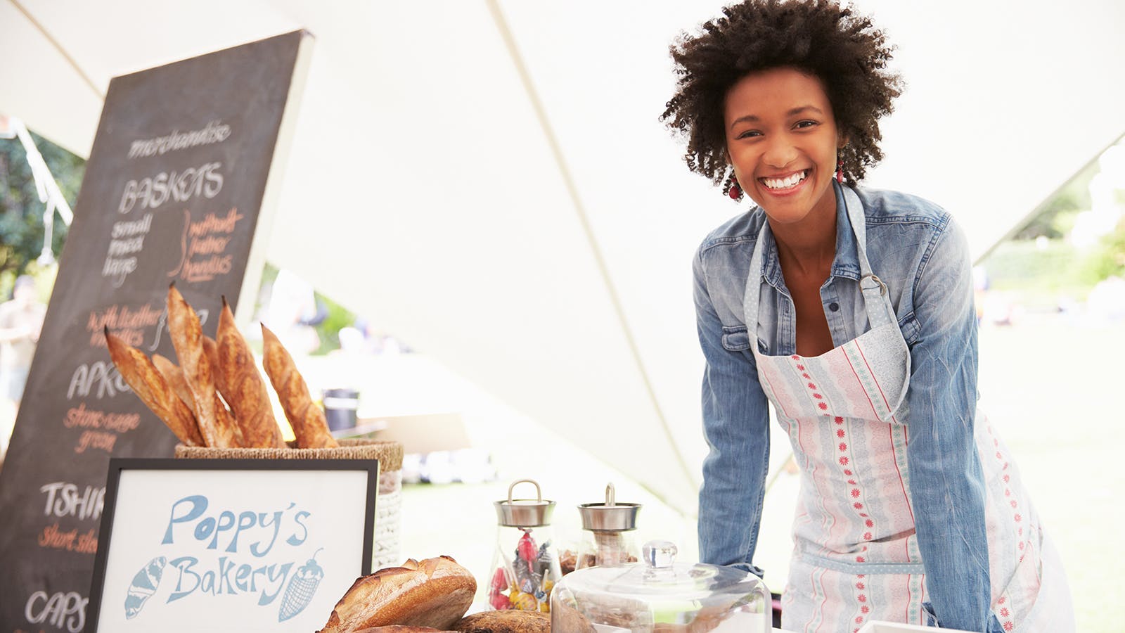 A smiling woman wearing an apron stands behind a table at an outdoor bakery stall with a sign reading Poppy’s Bakery, displaying baguettes and pastries, with a chalkboard menu in the background.