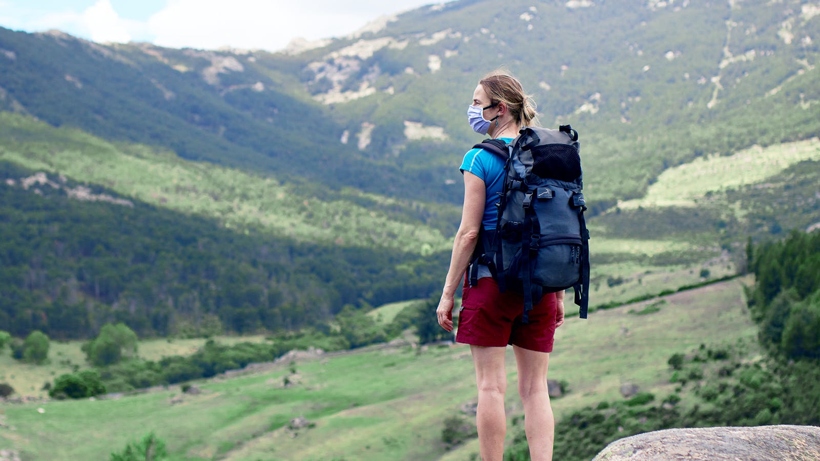 A woman wearing a face mask, blue shirt, and red shorts stands on a rock with a large backpack, looking out over a scenic green valley and forested mountains.