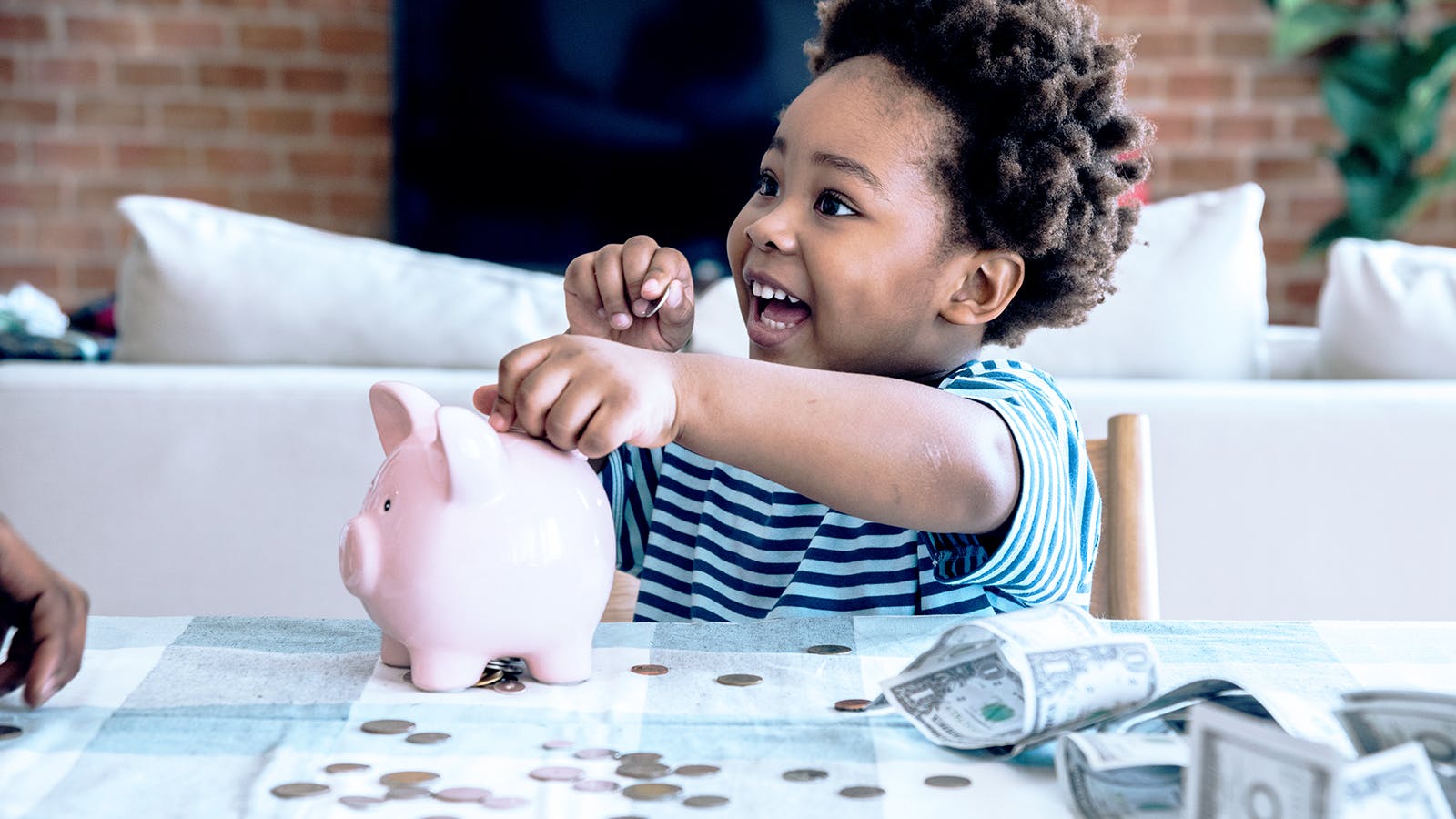 Smiling child in a striped shirt sits at a table, putting coins into a pink piggy bank, with coins and dollar bills scattered on the table in front of them.
