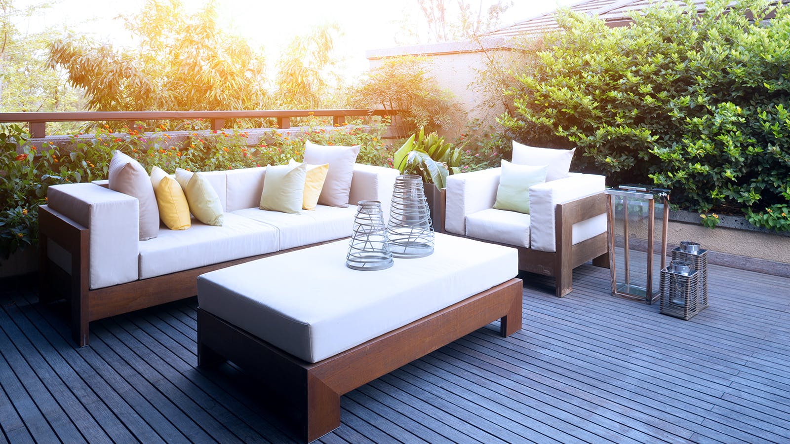 Outdoor patio with modern wooden furniture, including a sofa, armchair, and ottoman with white cushions and colorful pillows, on a wooden deck surrounded by lush greenery and soft sunlight.
