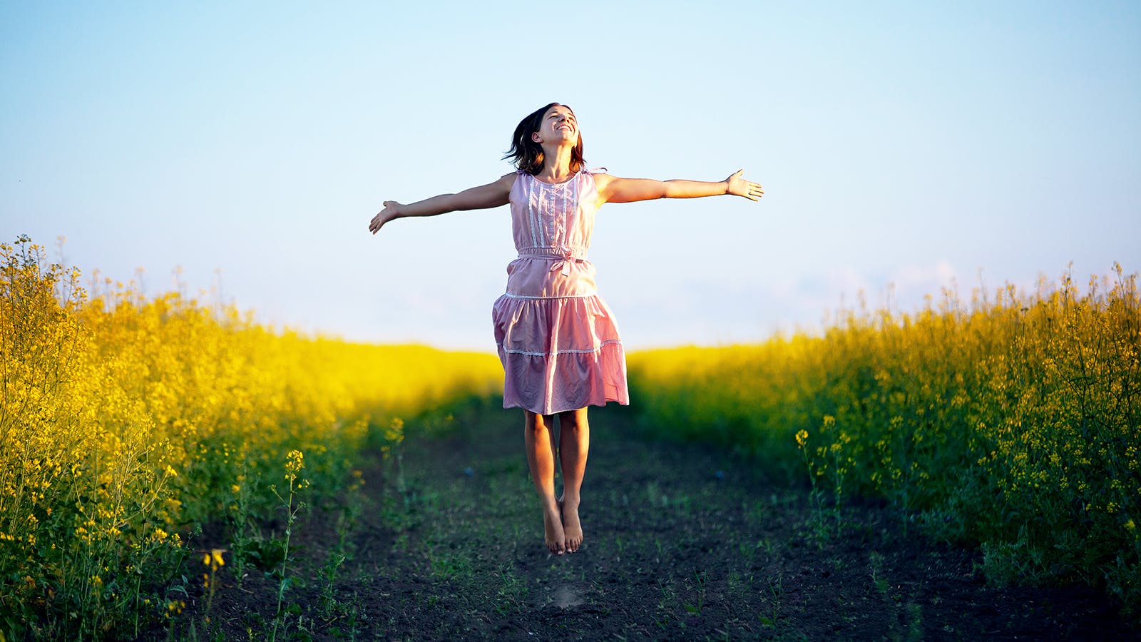 A woman in a pink dress joyfully jumps with outstretched arms in the middle of a vibrant yellow flower field under a clear blue sky.