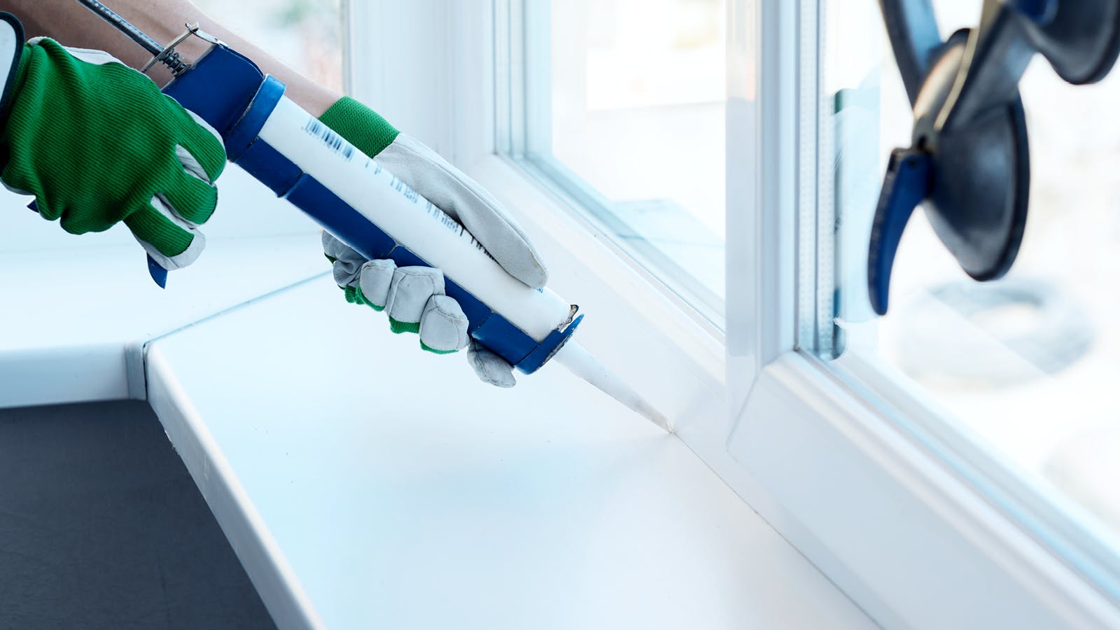 Person wearing green gloves using a caulking gun to seal the edge of a white window frame, applying white sealant along the seam between the window and the windowsill.