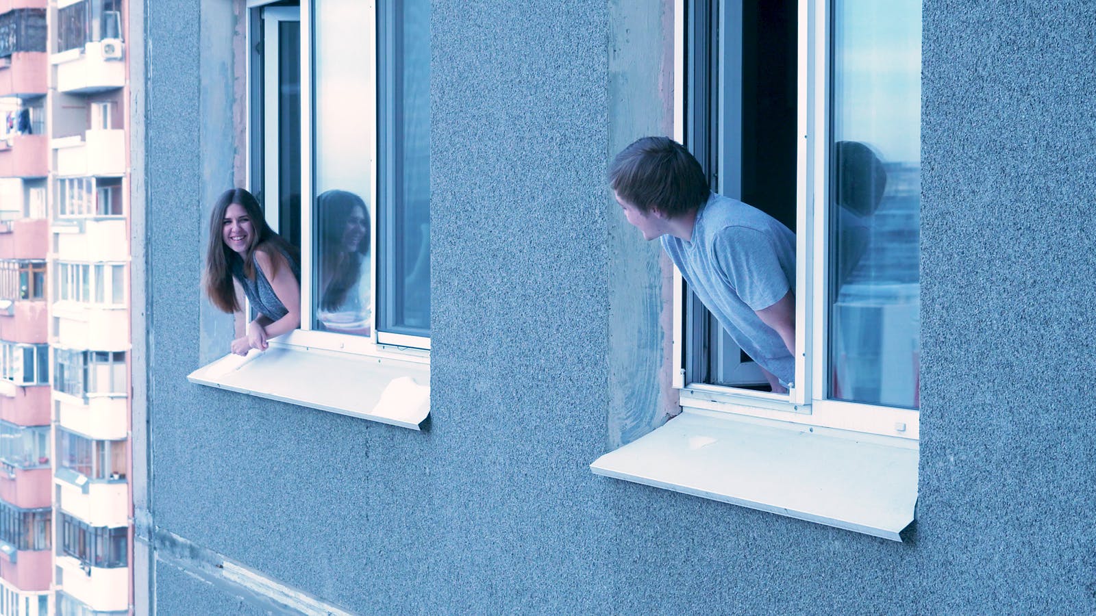 A young woman and a young man lean out of separate windows of a gray apartment building, smiling and looking at each other from a distance. The background shows another apartment building.