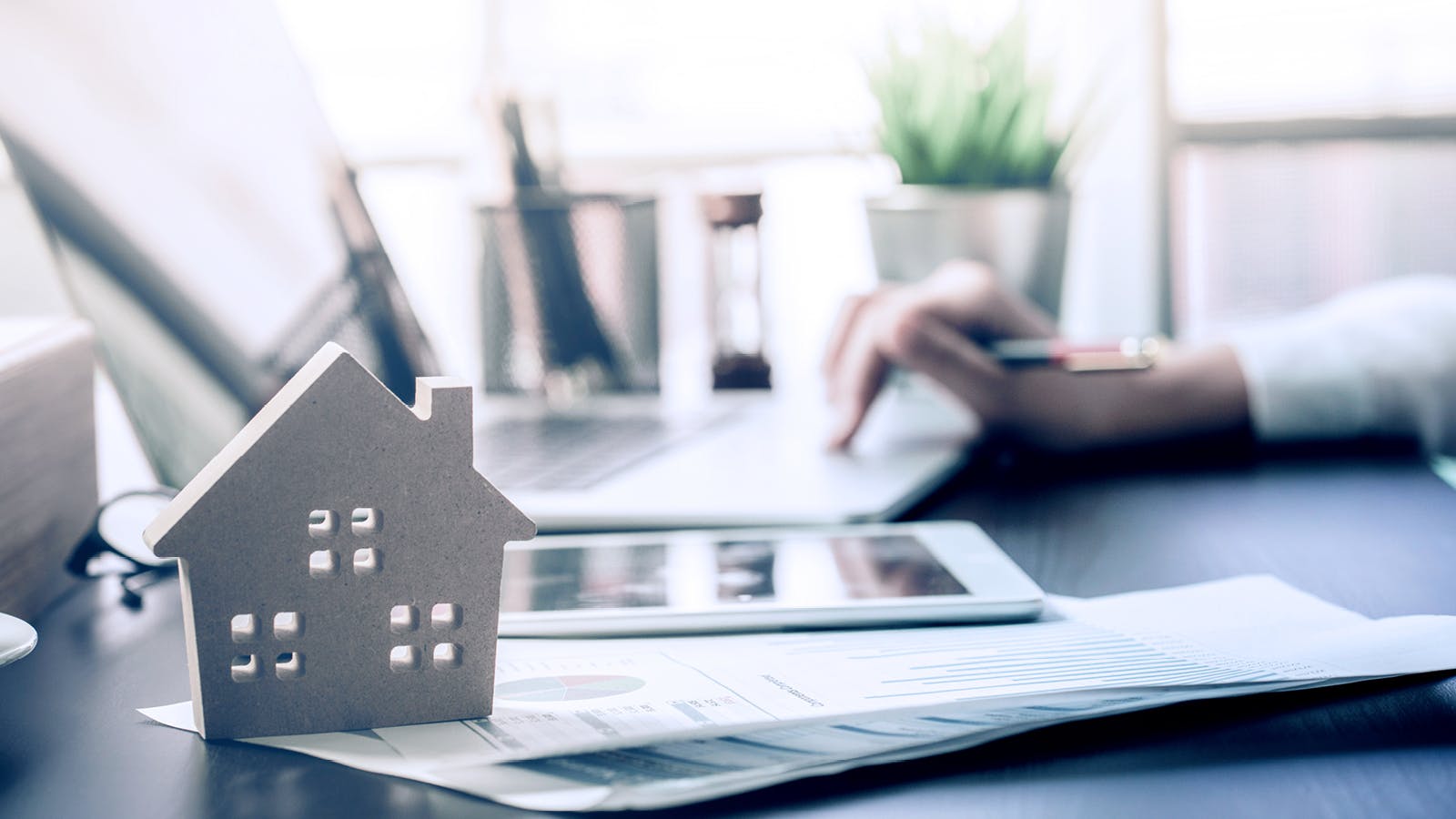 A small wooden house model sits on top of financial documents and charts next to a tablet, with a person typing on a laptop in the background at a desk.