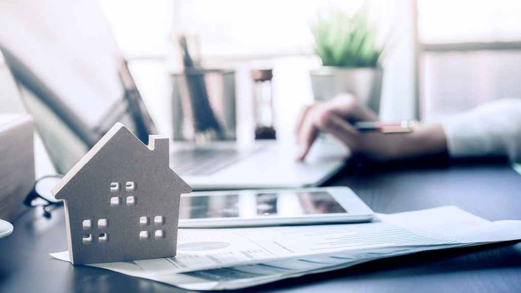 A small wooden house model sits on top of financial documents and charts next to a tablet, with a person typing on a laptop in the background at a desk.