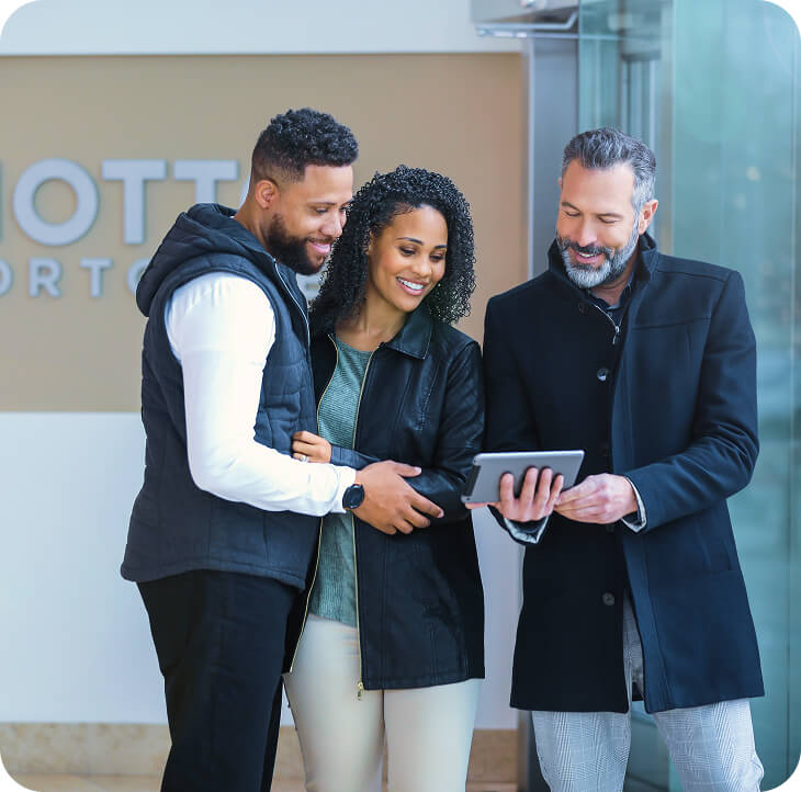 Three people standing indoors, smiling and looking at a tablet held by the man on the right. The group appears engaged and friendly, dressed in casual and business-casual winter clothing.