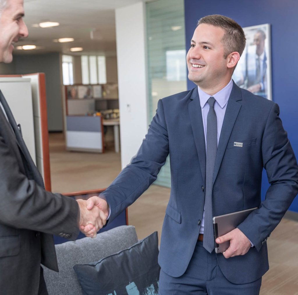 Corporate 1 Two men in business suits shaking hands in a modern office. One man is smiling and holding a tablet, while the other’s face is partially visible. Office furniture and cubicles are in the background.