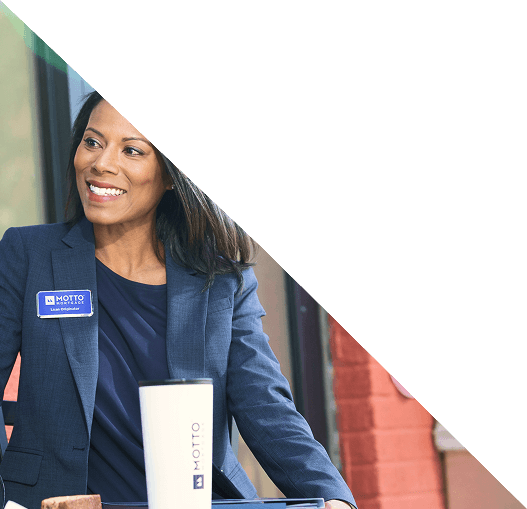 Hero (3) (1) A woman in a navy blazer with a MOTTO name tag smiles while sitting at a table with a white travel mug. The top right portion of the image is covered by a white triangle.