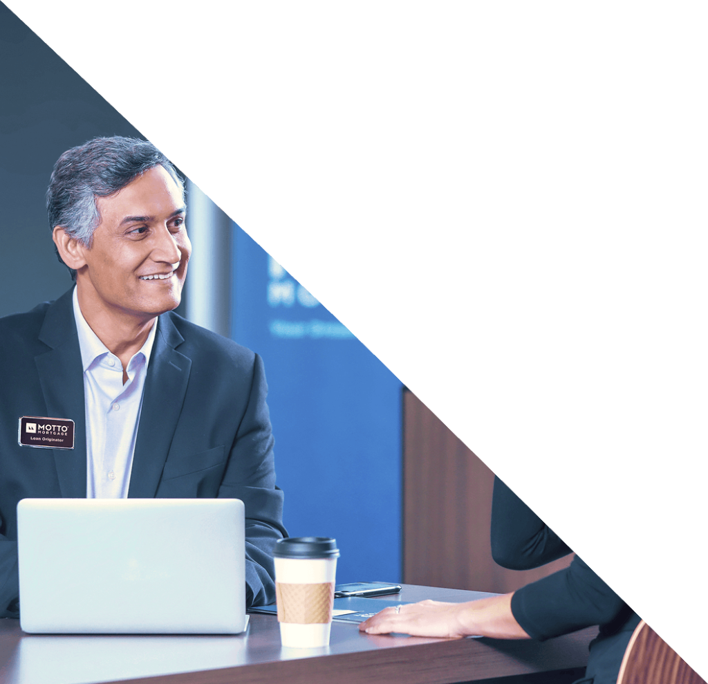 A man in a suit with a name badge sits at a table with a laptop and coffee cup, smiling and talking to another person, with a white diagonal area covering the top right half of the image.