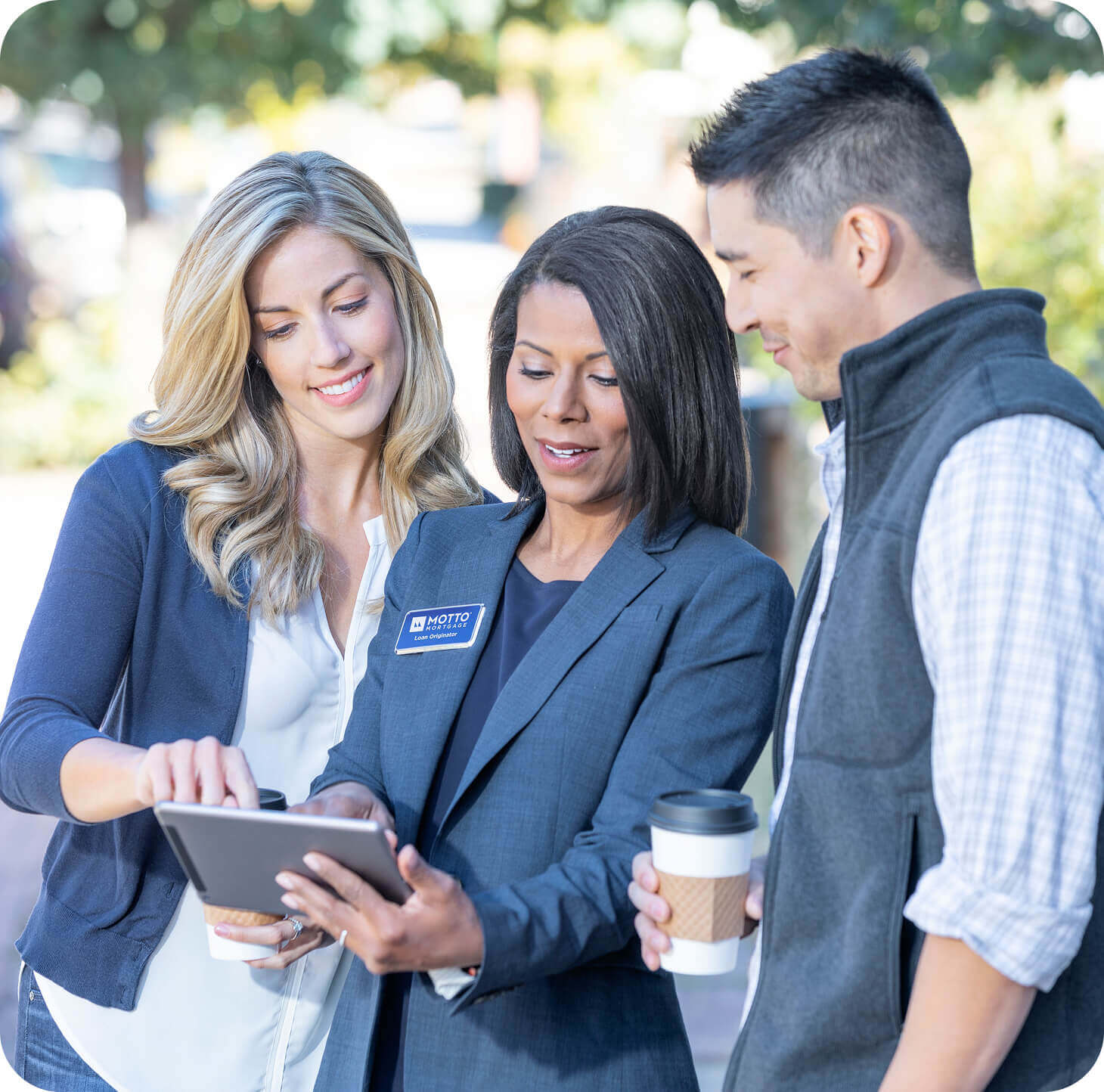 Image (2) (1) (1) Three people stand outdoors, smiling and looking at a tablet. The woman in the middle wears a blazer and name badge, and the other two hold coffee cups while engaging in the conversation. Trees and buildings are blurred in the background.