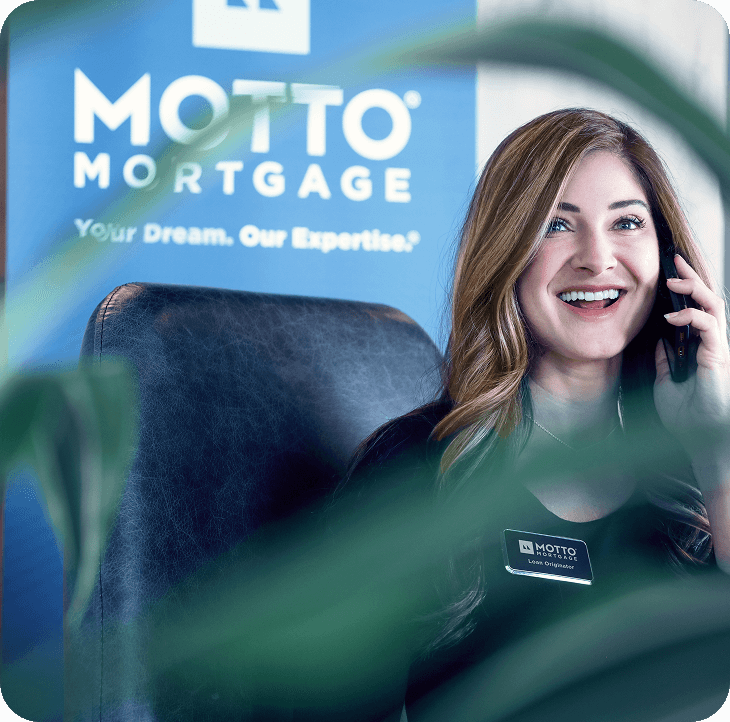 A woman with long brown hair sits at a desk, smiling while talking on the phone. A Motto Mortgage banner with the slogan Your Dream. Our Expertise. is visible in the background. Green leaves are in the foreground.