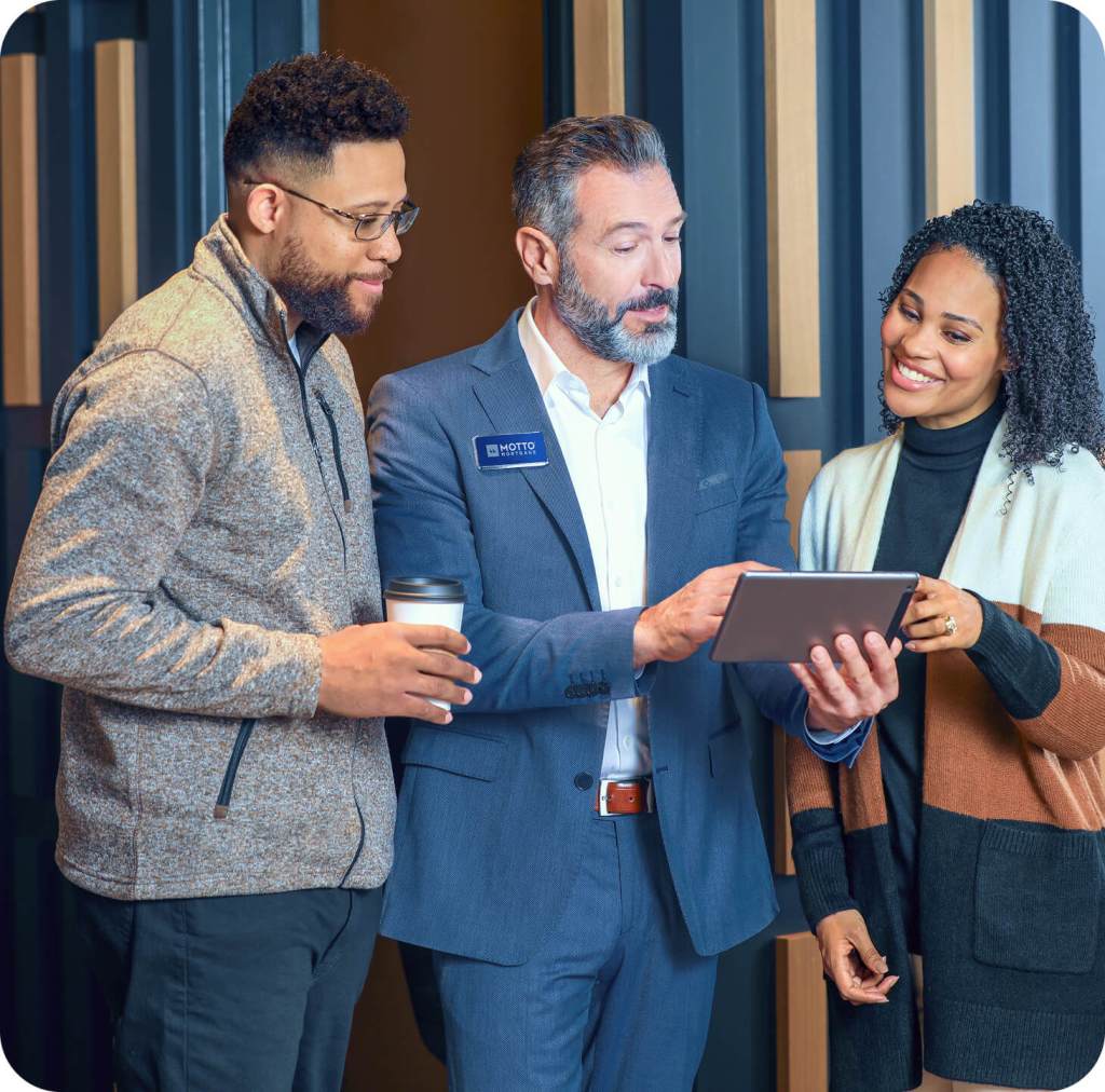 Mortgage (1) Three people stand together indoors, looking at a tablet held by a man in a blue suit with a name badge. One man holds a coffee cup, and the woman smiles as they all focus on the screen.