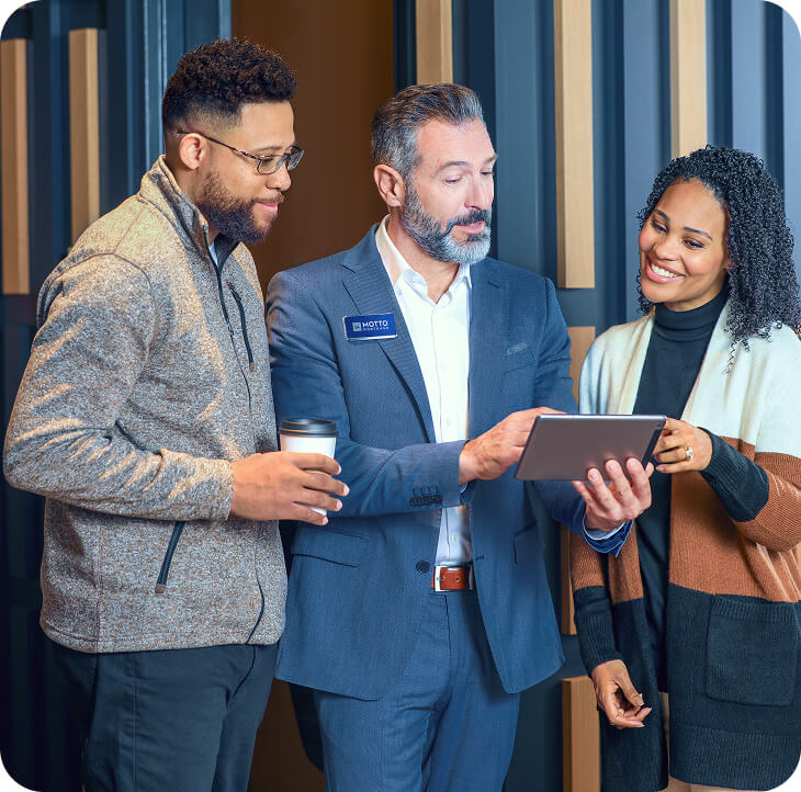 Three people stand together indoors, two casually dressed and one in a suit holding a tablet. The person in the suit points at the tablet while the others look on, smiling and engaged in conversation.