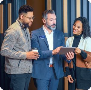 There’s More to a Mortgage Than the Rate (1) Three people stand together indoors, two casually dressed and one in a suit holding a tablet. The person in the suit points at the tablet while the others look on, smiling and engaged in conversation.