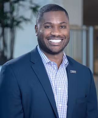 A man wearing a dark blue blazer and a checked dress shirt smiles at the camera. The background is softly blurred, showing indoor plants and a light-colored wall.