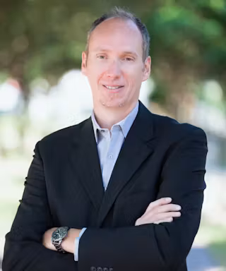 A man in a dark suit with a light shirt stands outdoors, smiling with arms crossed. Blurred green trees are in the background.