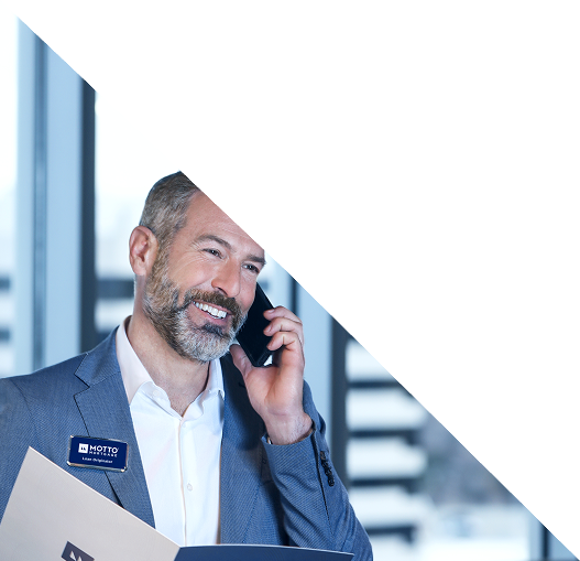 A smiling man in a suit holds papers and talks on a phone. He wears a name badge and stands in front of large windows with blurred city buildings in the background. The top of the image is covered by a white triangle.