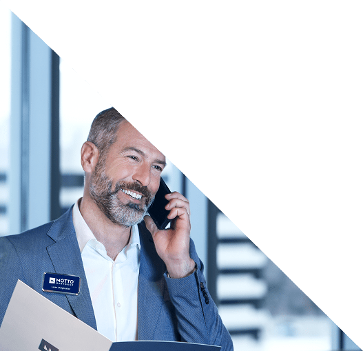 AboutHero A smiling man in a suit holds papers and talks on a phone. He wears a name badge and stands in front of large windows with blurred city buildings in the background. The top of the image is covered by a white triangle.