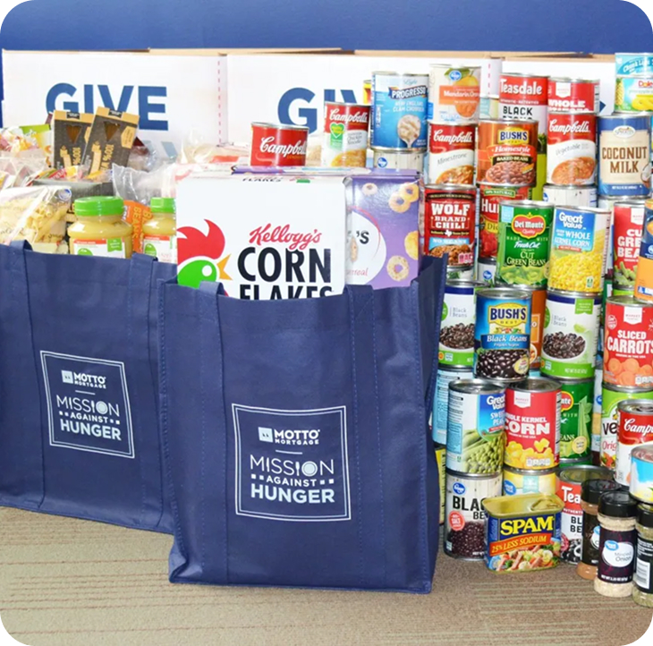 Community Blue tote bags labeled Mission Against Hunger are filled with food, sitting in front of stacks of canned goods, cereal, and other non-perishable items, with signs reading GIVE in the background.