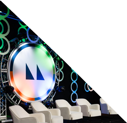 A modern stage setup with white chairs and a backdrop of colorful circular decorations, featuring a large circular logo with two upward-pointing triangles in the center. A white diagonal overlay covers the top right corner.