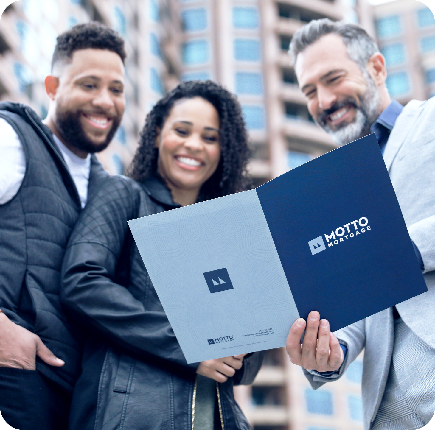 Three people smiling and looking at a folder labeled Motto Mortgage, standing outdoors in front of a modern building.