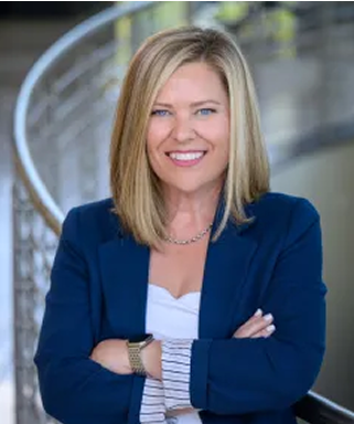A woman with straight blonde hair, wearing a blue blazer and white top, stands with arms crossed and smiles confidently. The background is blurred, showing a curved railing and modern indoor setting.