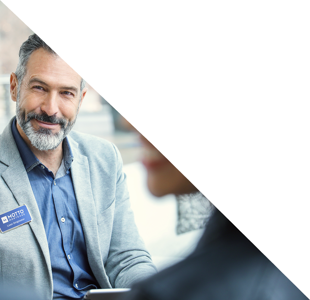 A man with gray hair and a beard, wearing a light blue blazer and a name tag, smiles while talking to another person. The top right half of the image is covered by a white triangle.