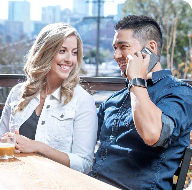 Rectangle (1) A woman and a man sit together at an outdoor table. The woman smiles while looking at the man, who is talking on a smartphone and smiling. A cityscape and trees are visible in the background.