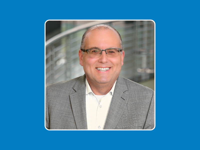 A smiling man wearing glasses, a grey blazer, and a white shirt poses in front of a blurred background with metal railings. The image is framed with a white border and a blue background.
