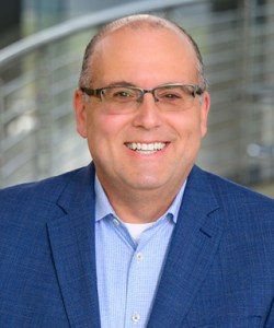 Smiling man with glasses wearing a blue suit jacket and light blue shirt, standing in front of a blurred background with metal railings.