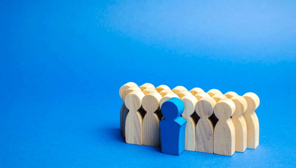 A group of wooden figurines stands together, with one distinct blue figure positioned in front of the group, against a solid blue background.